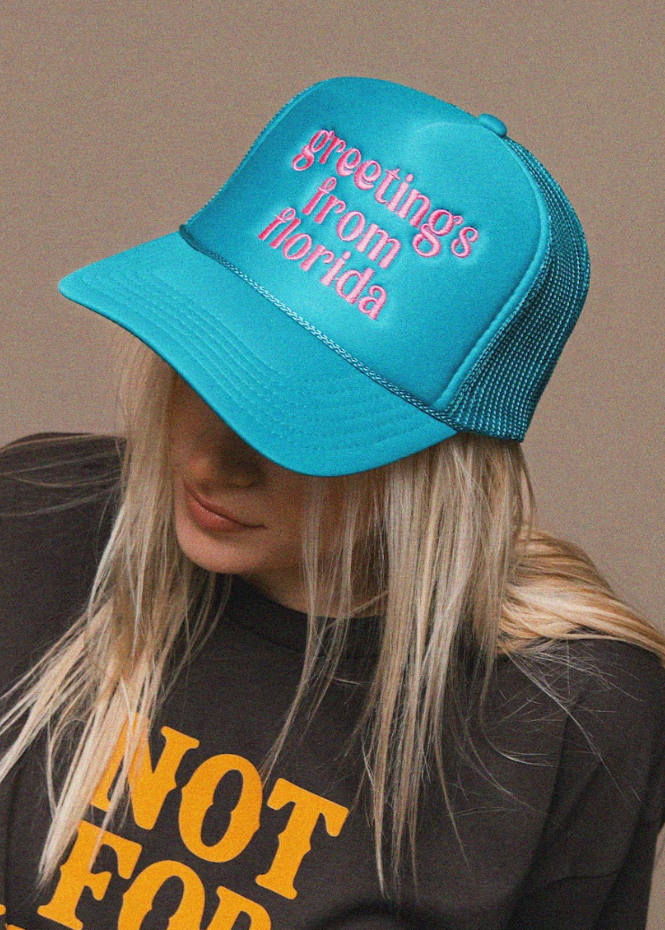 Blue cap with 'greetings from florida' text worn by a person, against a beige background