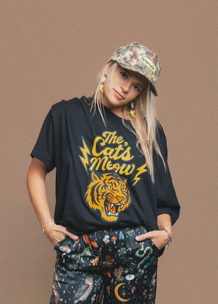 Person wearing a black t-shirt with a tiger graphic and 'The Cat's Meow' text, standing against a brown background.