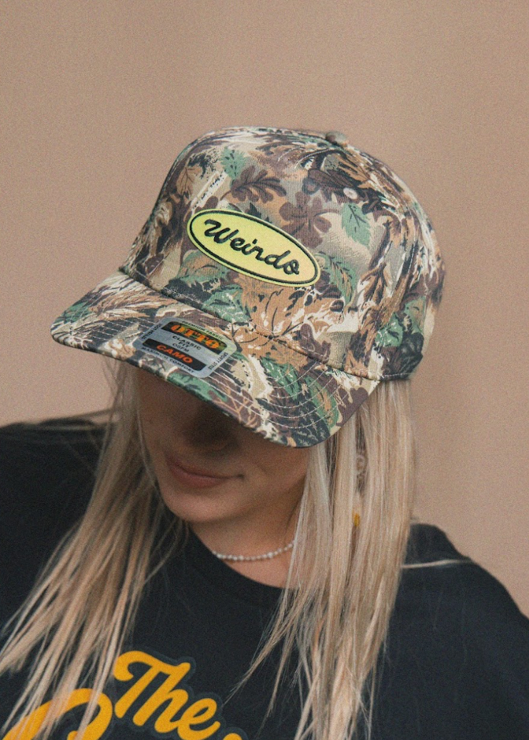 Person wearing a camouflage cap with 'Weinb' logo on a beige background