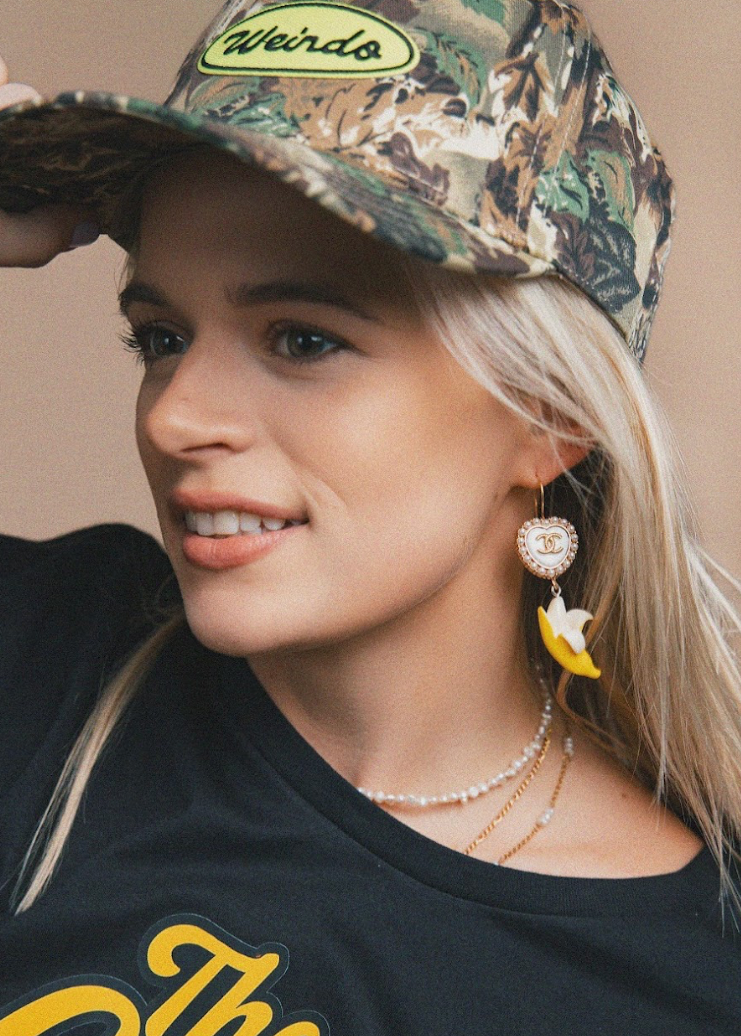 Woman wearing a camouflage cap with 'Weirdo' text and a black shirt with yellow text, against a beige background.