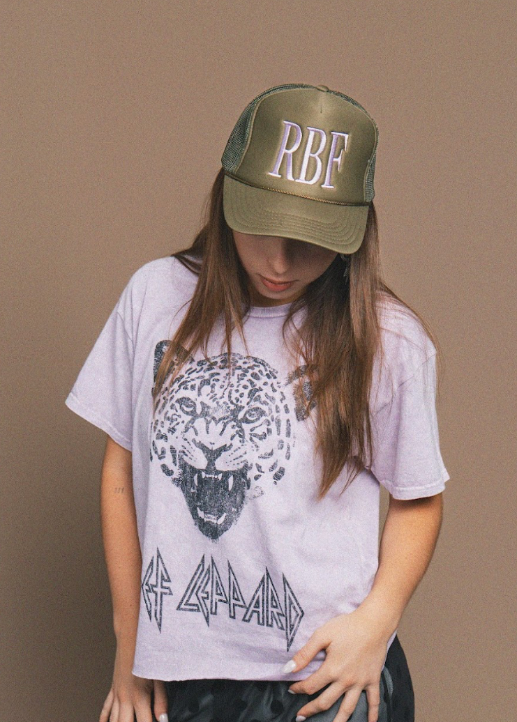 Person wearing a white t-shirt with a graphic design and an olive green cap with 'RBF' on a brown background