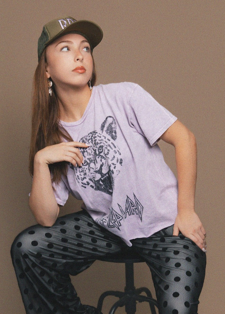 Woman wearing a gray t-shirt with a graphic design and black polka dot pants, sitting on a chair against a brown background.
