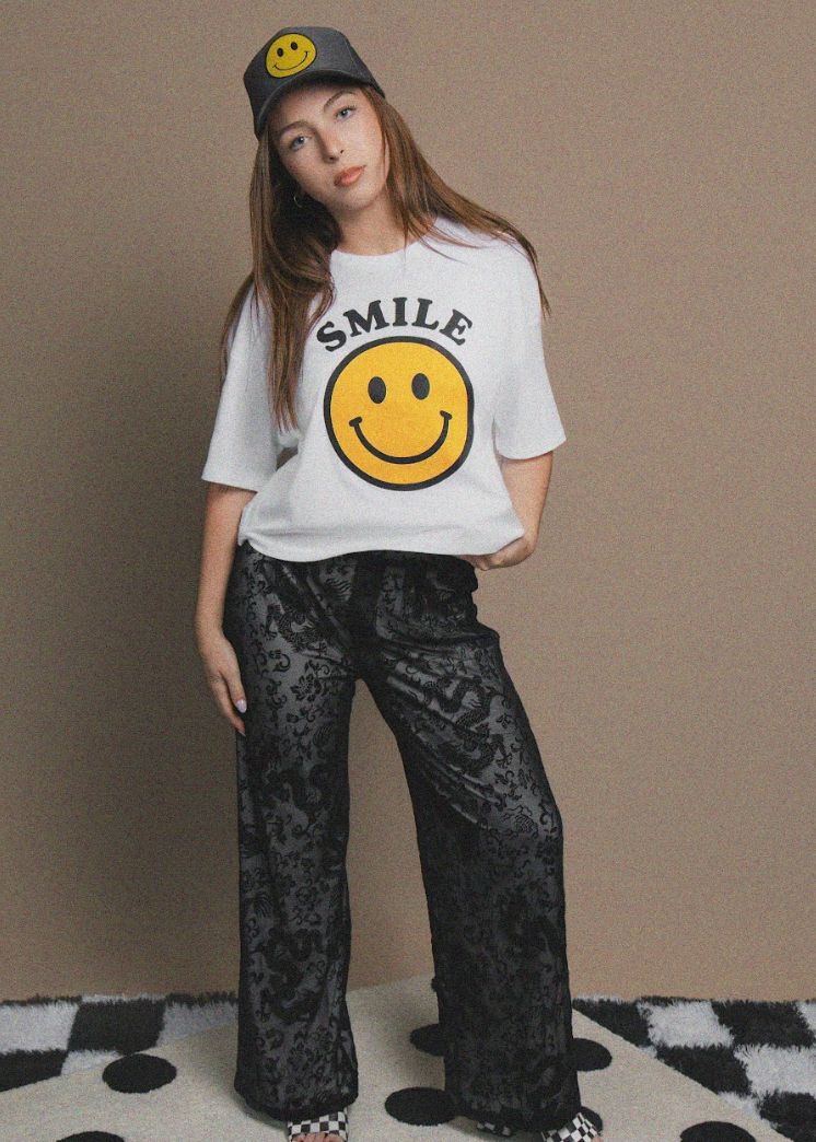 Person wearing a white t-shirt with a smiley face and 'SMILE' text, black pants, and a gray cap with a smiley face on a beige background.