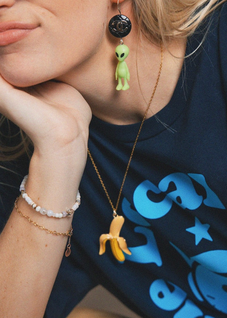 Person wearing a necklace with a banana pendant and alien earrings, against a dark blue shirt with cartoon designs.