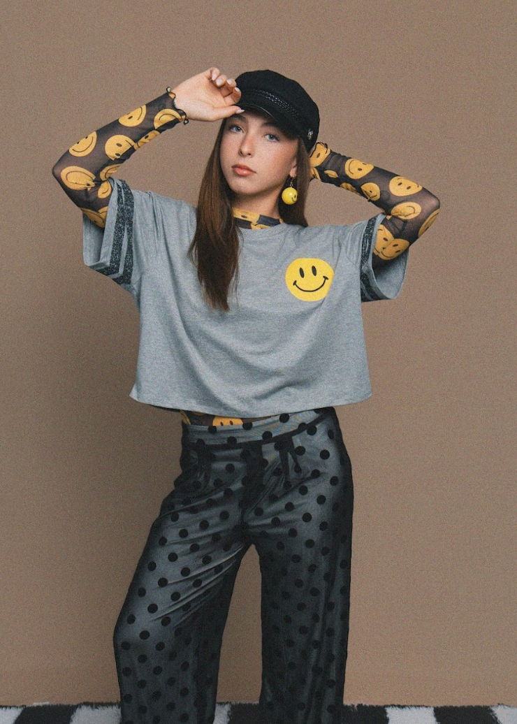 A model posing in a gray cropped t-shirt with a yellow smiley face graphic, matching smiley-print long sleeves, black polka-dot pants, a black cap, and yellow smiley-face earrings, set against a solid tan background.