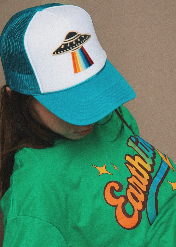 Person wearing a teal and white cap with a UFO design, standing against a beige background.