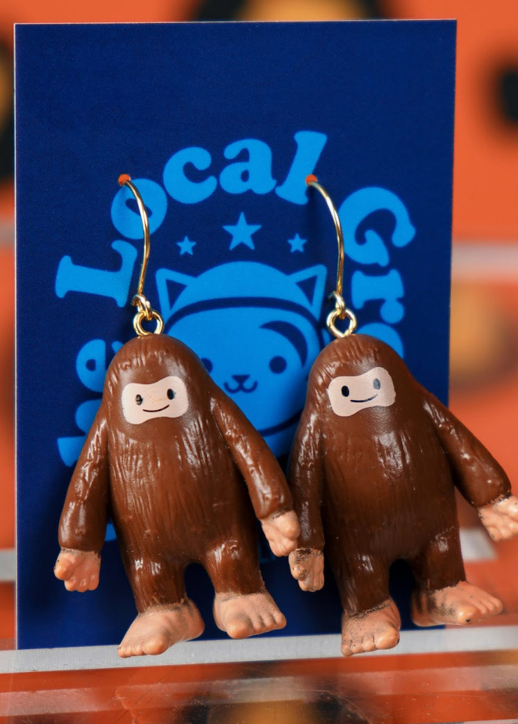 Brown cartoon-like creature earrings on a blue card with a logo.