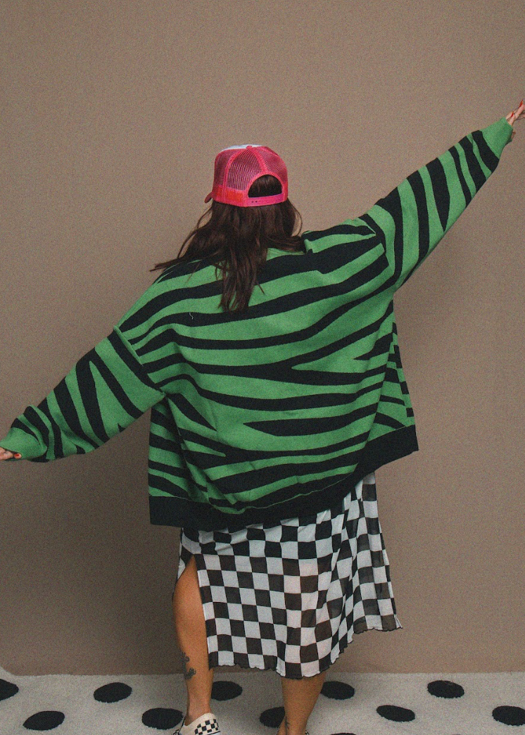 Person wearing a green and black zebra-striped sweater and pink cap, standing against a plain wall.
