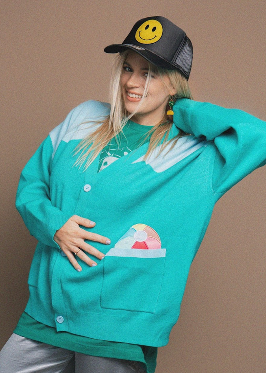 Person wearing a teal cardigan with a colorful design and a smiley face cap on a brown background