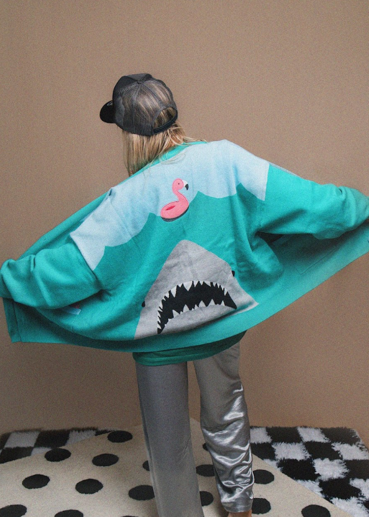 Person wearing a shark-themed hoodie with a pink flamingo on a checkered floor.