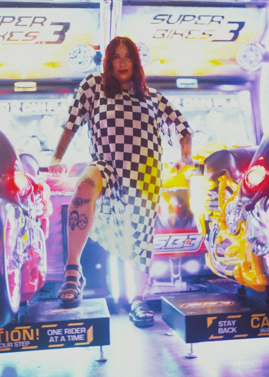 Person in a checkered dress standing in front of a 'Super Bikes 3' arcade machine.