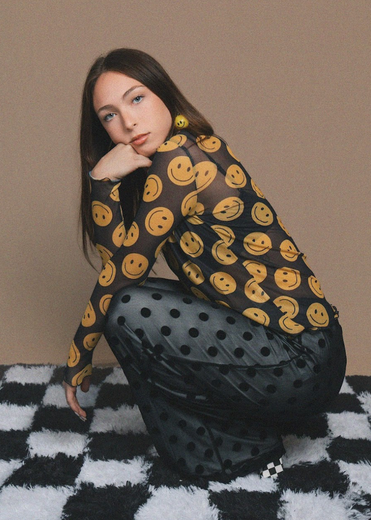 Woman wearing a sheer top with smiley face patterns on a checkered floor.