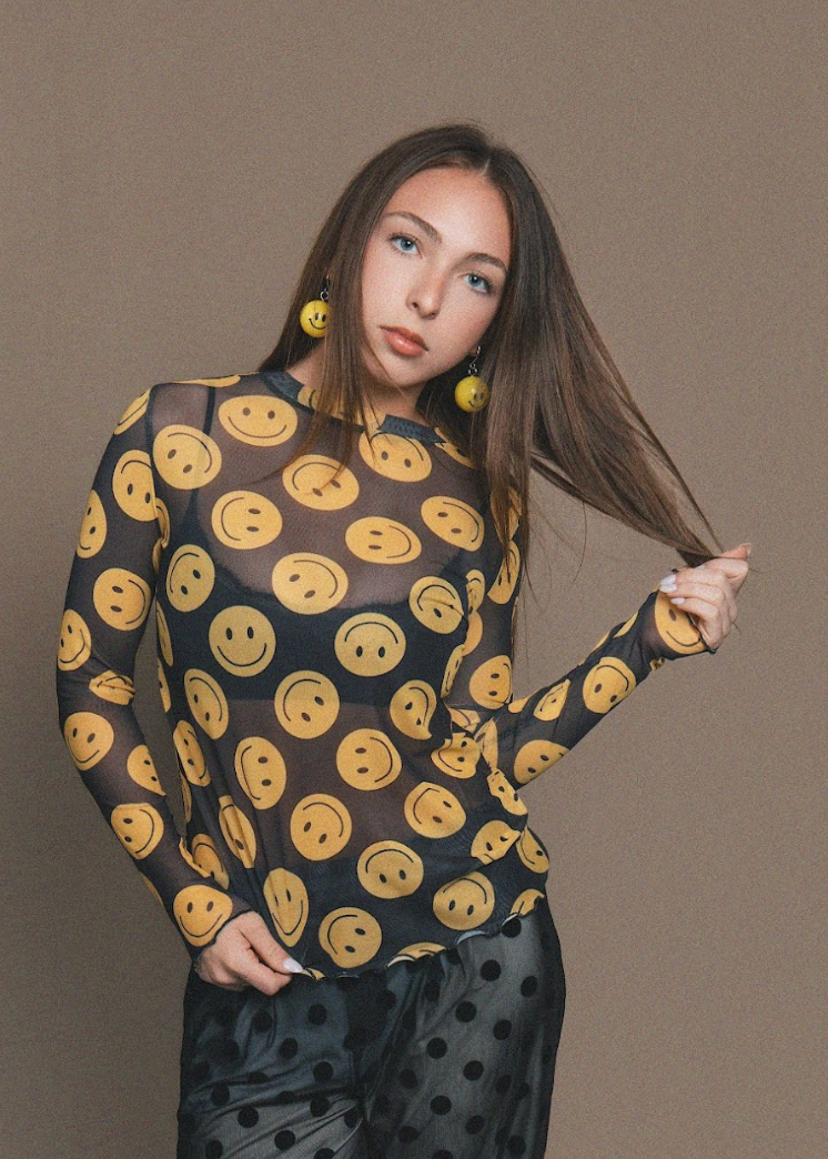 Woman wearing a sheer black top with yellow smiley face patterns against a brown background
