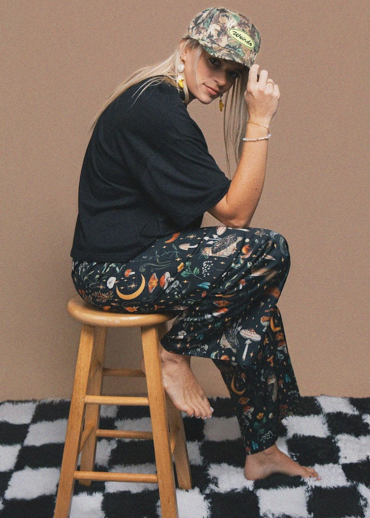 Person sitting on a stool wearing a black t-shirt and patterned pants with a camouflage cap.