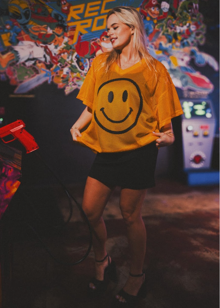 Person wearing a yellow smiley face shirt in front of a colorful arcade machine.