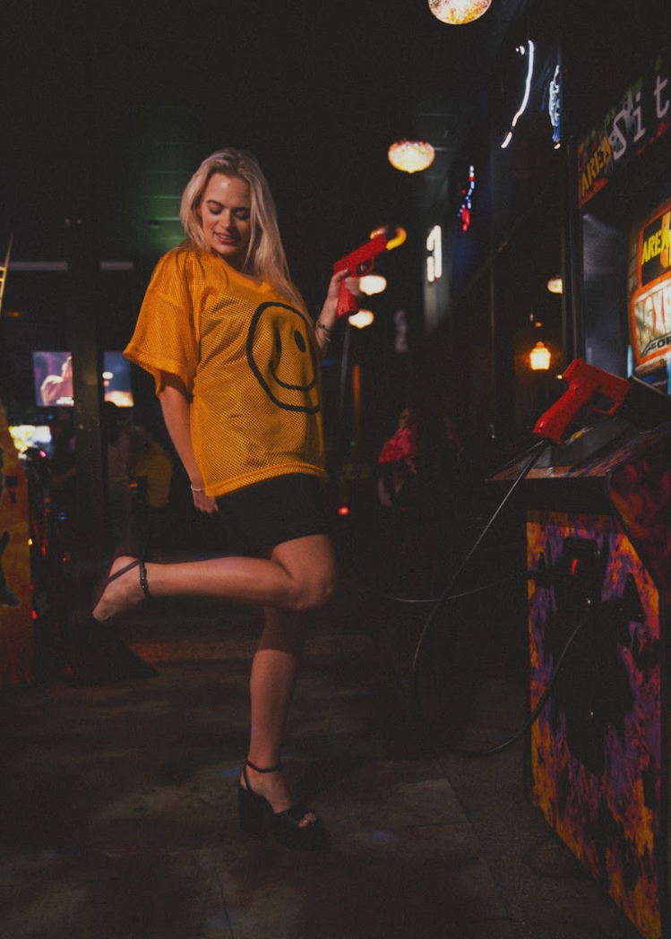 Person in a yellow shirt with a smiley face design in a dark, arcade-like setting.