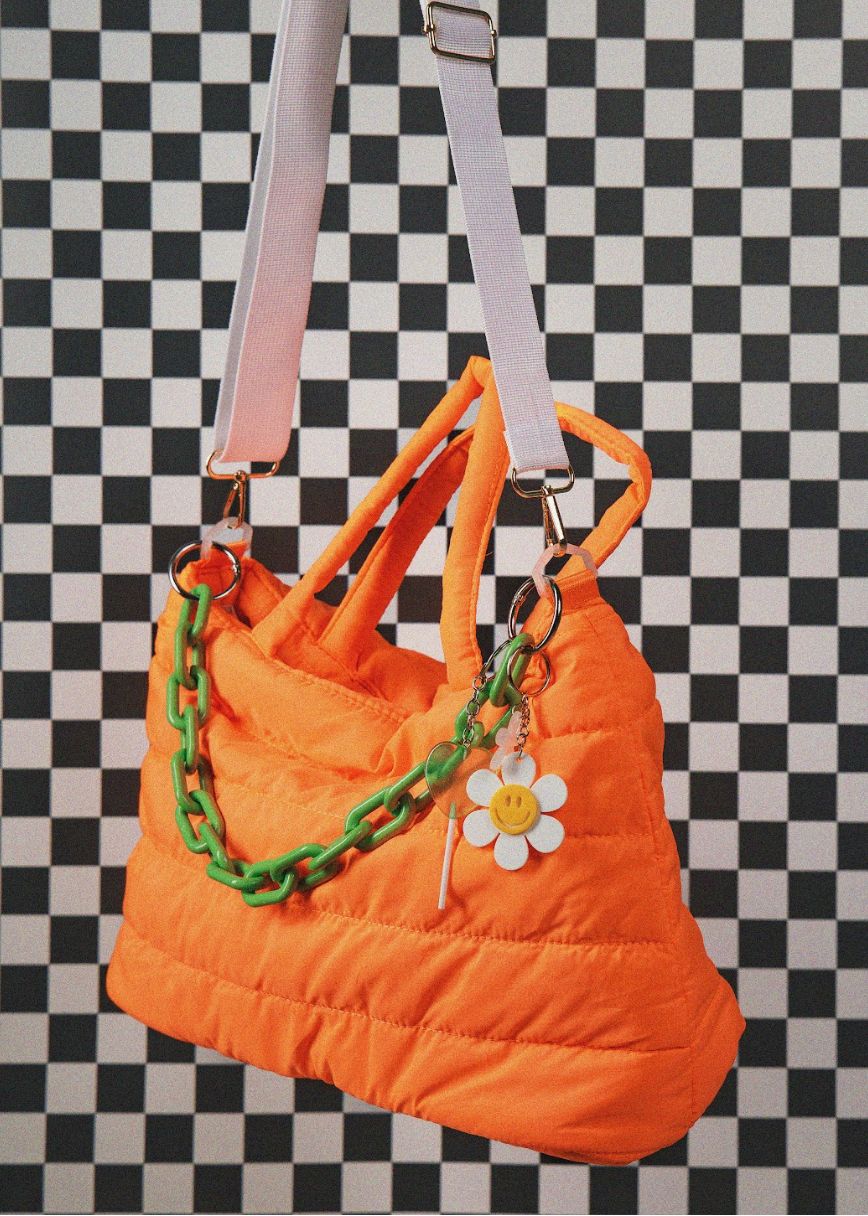An orange puffer-style shoulder bag with a white shoulder strap, green chain detail, and a white daisy charm with a yellow smiley face, shown against a black-and-white checkered background.