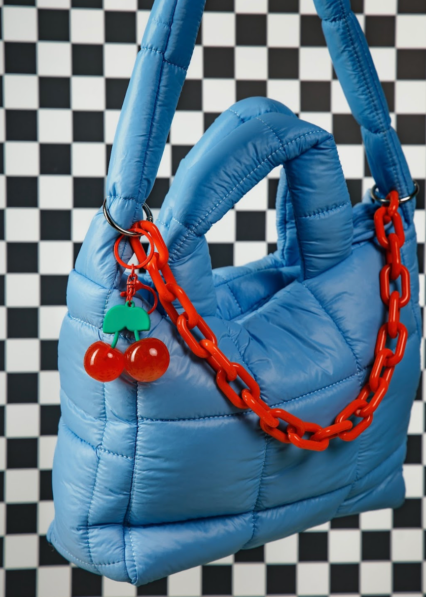 A light blue puffer handbag with a chunky red chain handle and a cherry-shaped charm, set against a black and white checkered background.