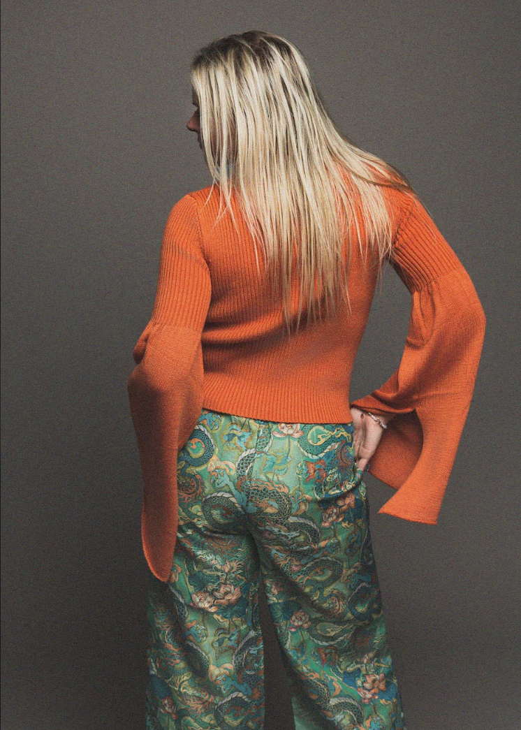 Person wearing an orange sweater and patterned pants against a gray background