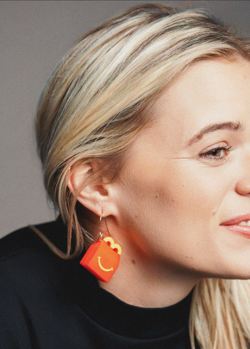 Close-up of a person wearing orange earrings with smiley face designs against a neutral background