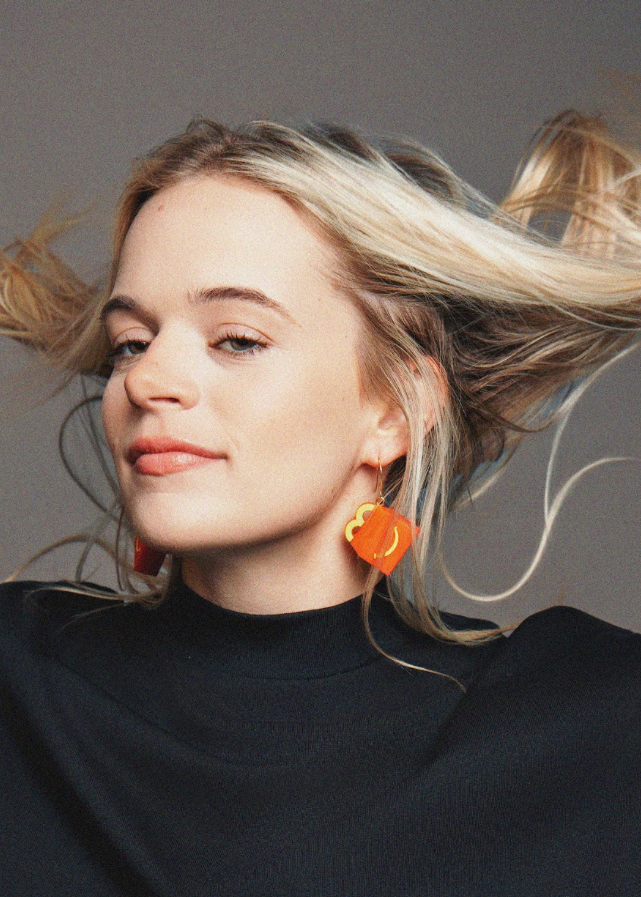 Woman with blonde hair and orange earrings against a gray background