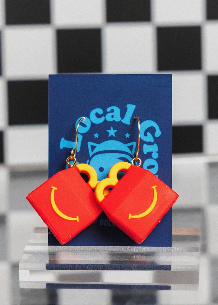 Red earrings with yellow smiley faces on a blue card with 'Local Group' branding against a checkered background.