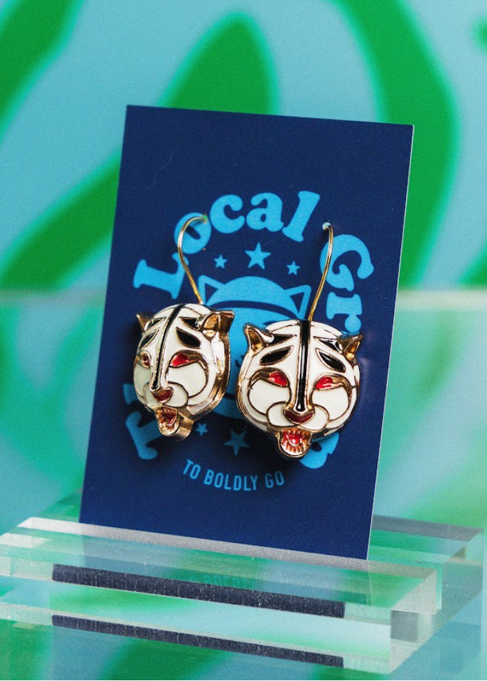 Pair of cat-shaped earrings on a blue card with 'Local Group' branding against a green and blue abstract background.