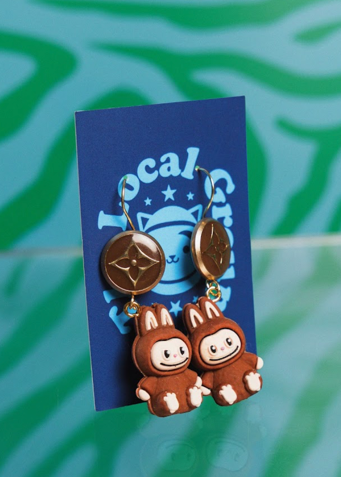 Pair of brown cartoon-like earrings on a blue card with 'Local Group' branding against a green patterned background.