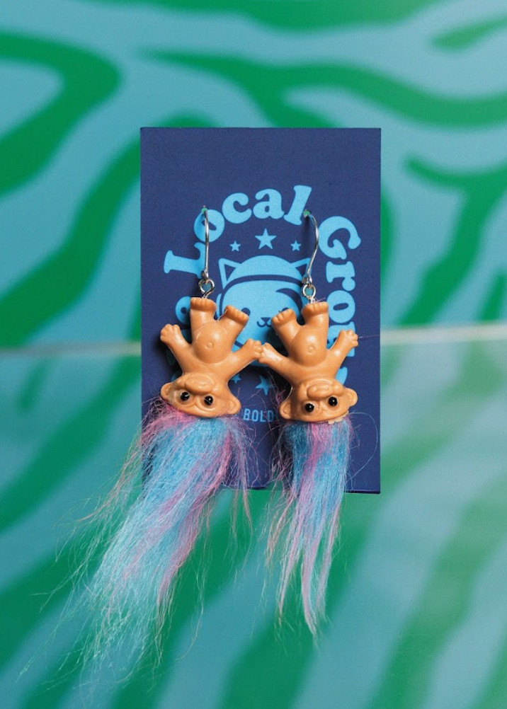Earrings with troll design and colorful tassels on a blue card with 'Local Group' text against a green patterned background.