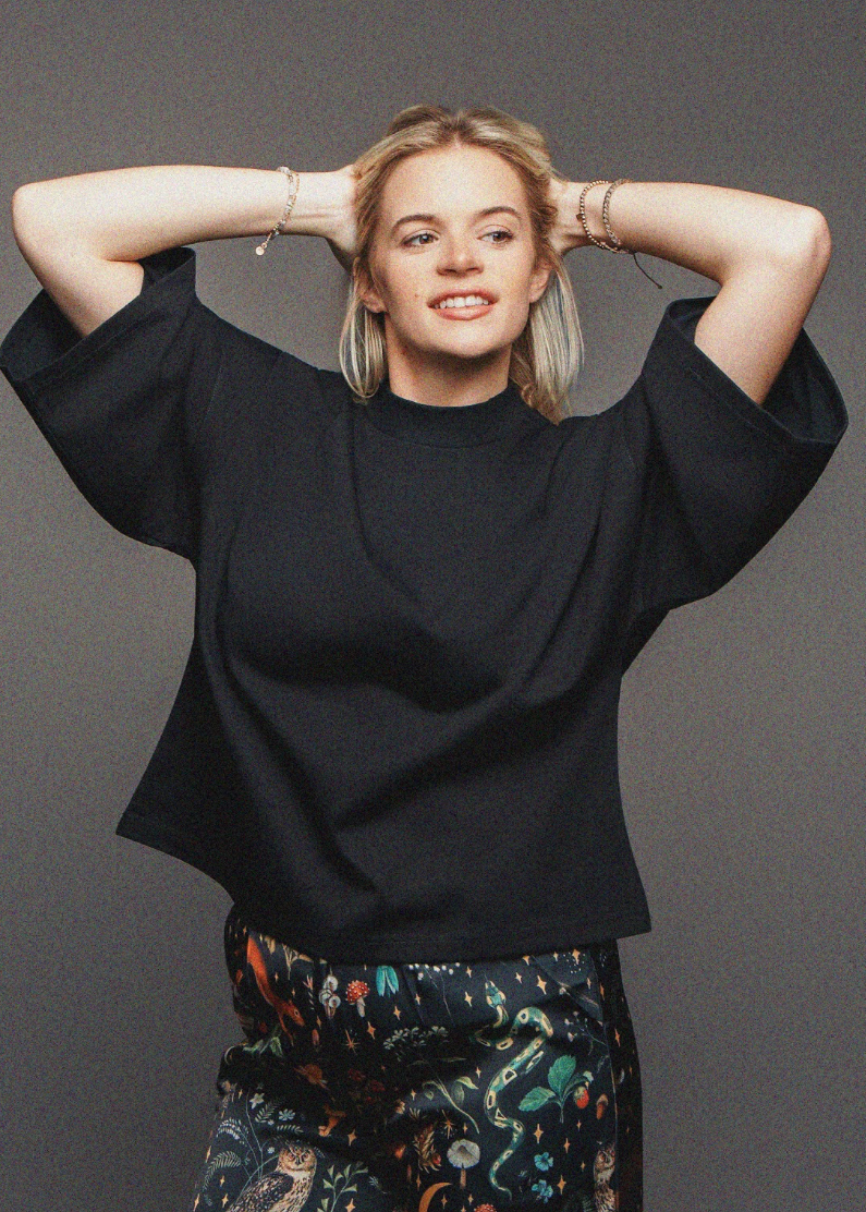 Person wearing a black top and patterned pants with hands behind head on a gray background