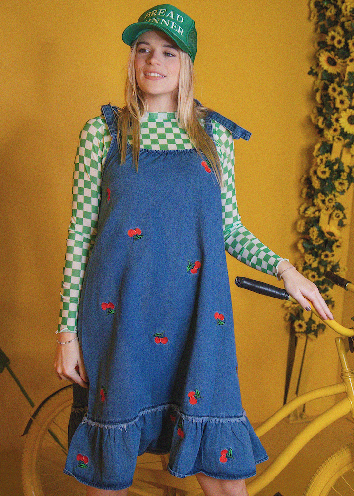 Person wearing a blue denim dress with red embroidery and a green cap, standing against a yellow wall.
