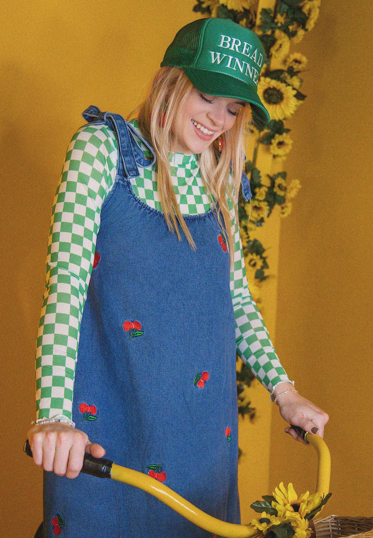 Person wearing a blue dress with red floral patterns and a green cap with text, standing against a yellow background with sunflowers.