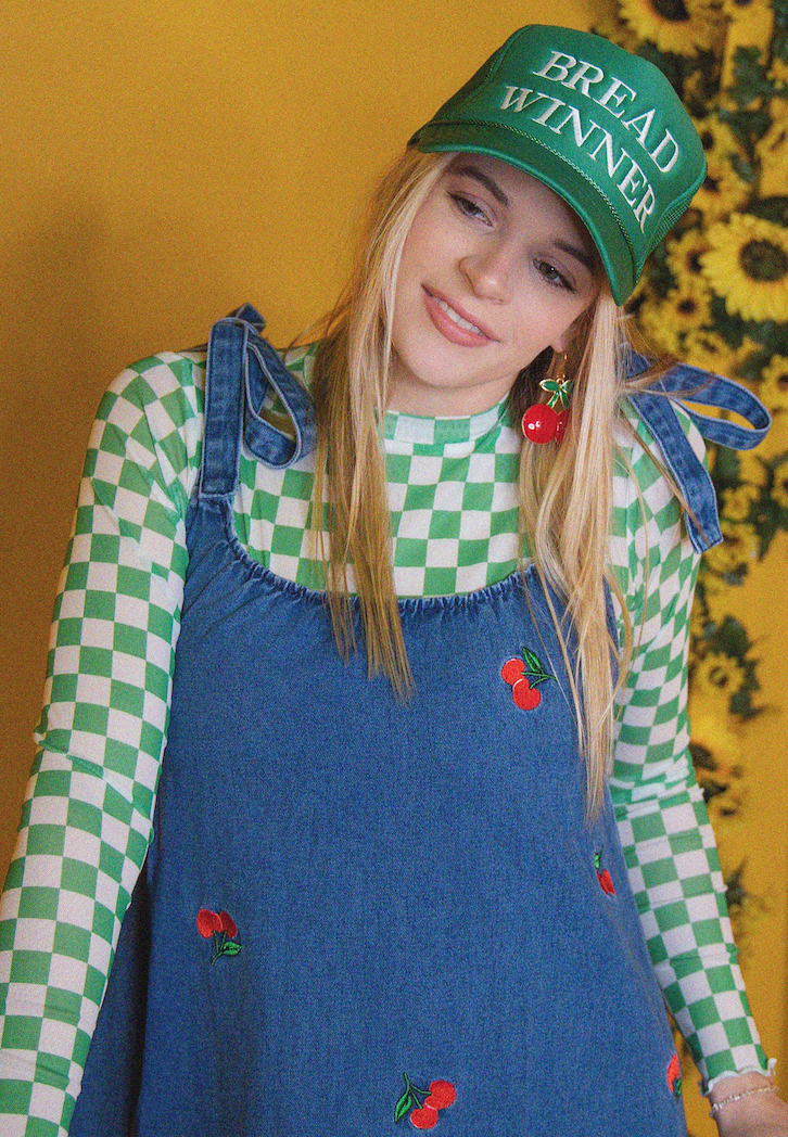 Person wearing a green 'Bread Winner' cap, blue dress with red cherries, and checkered shirt against a yellow background.