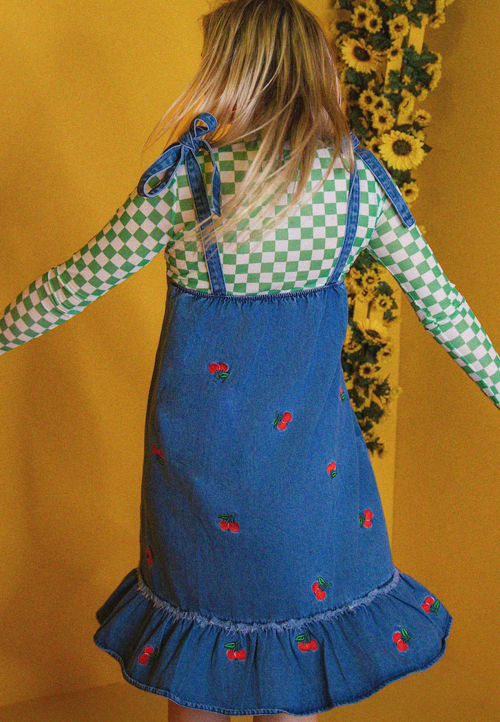 Person wearing a green checkered shirt and blue denim dress with red embroidery on a yellow background