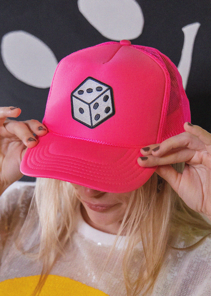 Person holding a pink cap with a dice design in front of their face against a black background with white shapes.