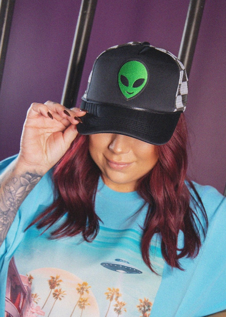 Person wearing a black cap with an alien face design, holding it over their eyes against a purple background.
