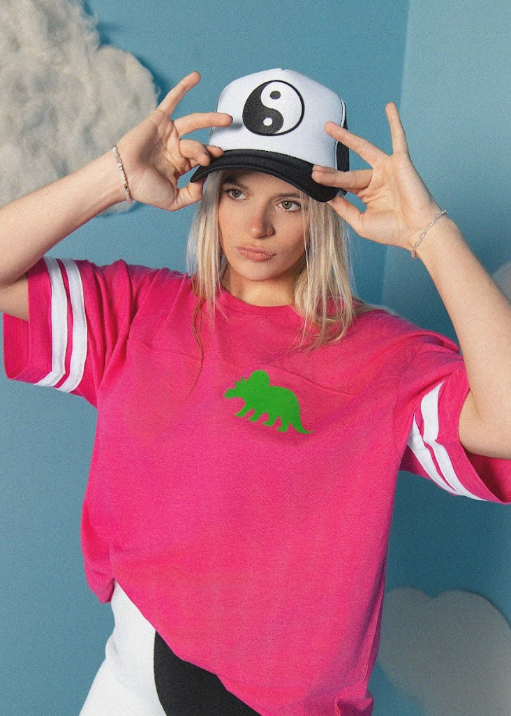 Person wearing a pink t-shirt with a green dinosaur graphic and a cap with a yin-yang symbol.