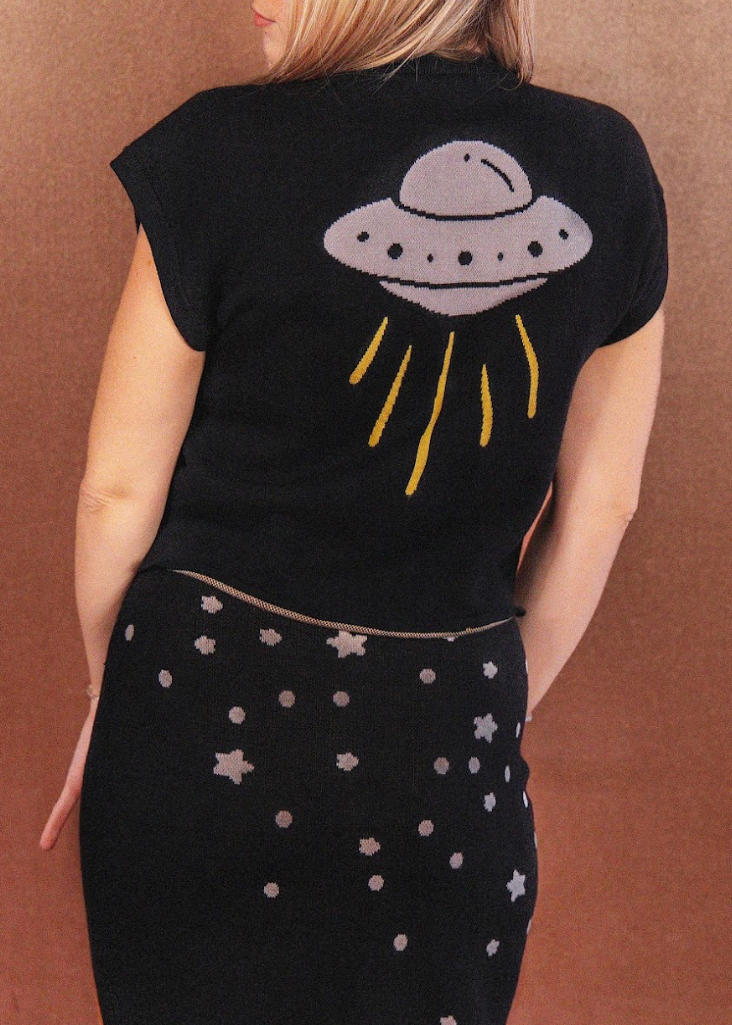 Person wearing a black t-shirt with a UFO design on the back, standing against a brown background.