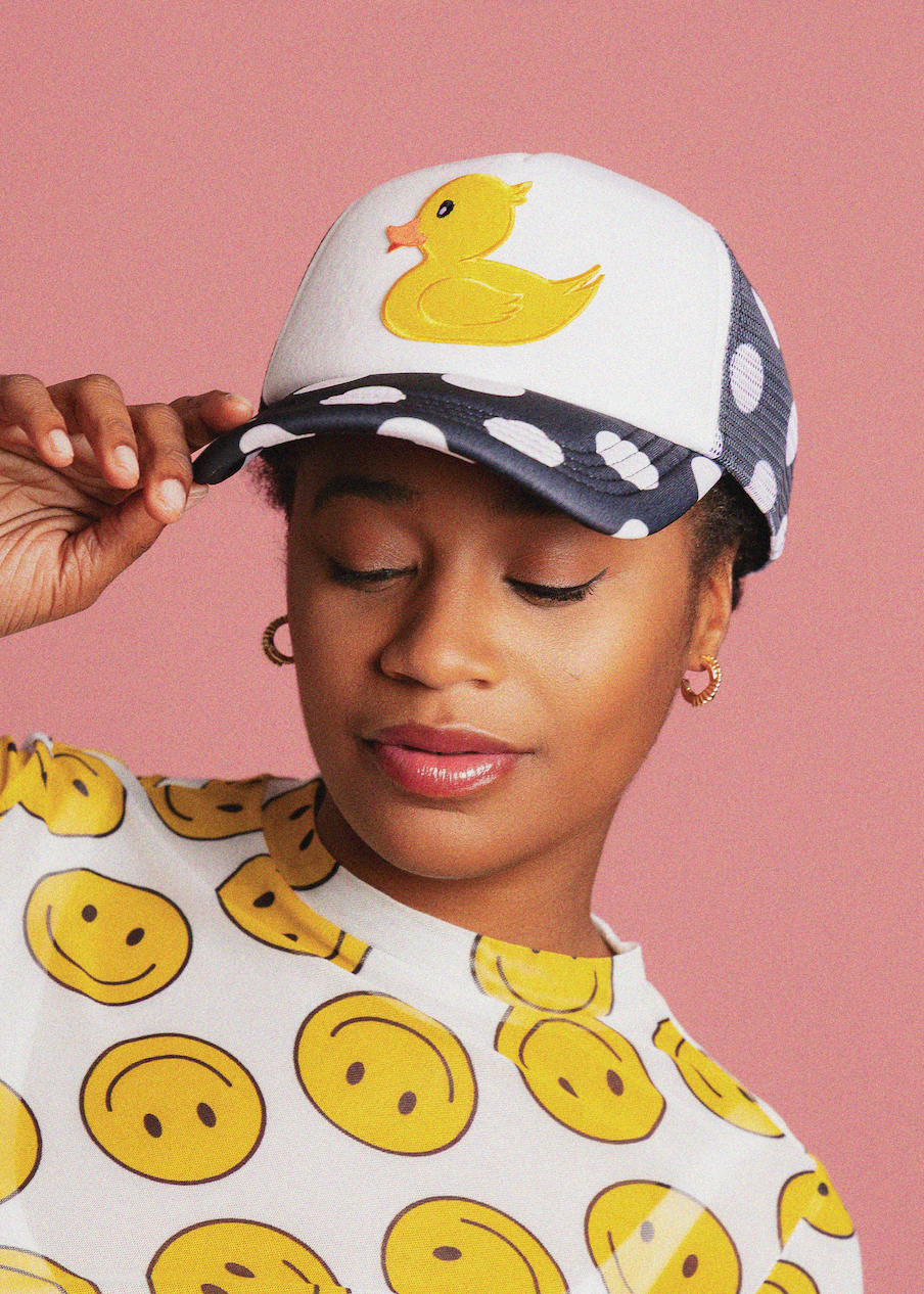Person wearing a cap with a duck design and a shirt with smiley face patterns against a pink background