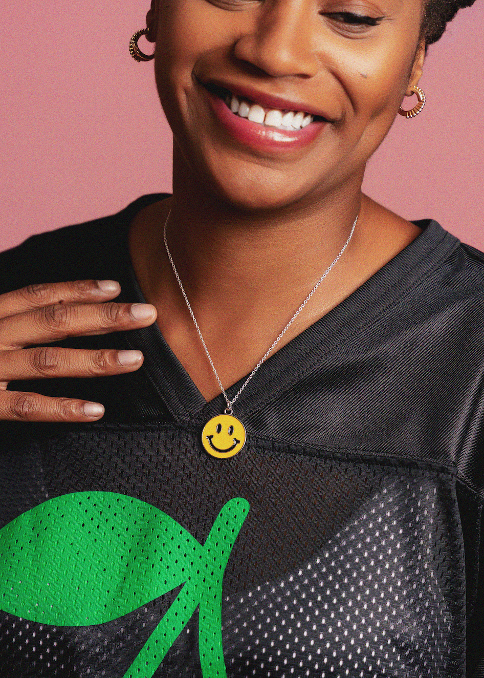 Person wearing a black sports jersey with a green logo and a yellow smiley face necklace on a pink background