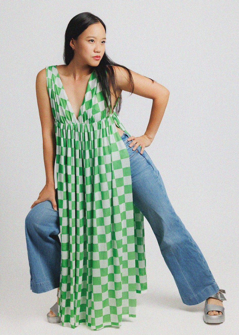 A woman modeling a green and white checkered sleeveless maxi dress with a deep V-neckline, styled with wide-leg blue jeans and silver platform sandals against a light gray studio background.