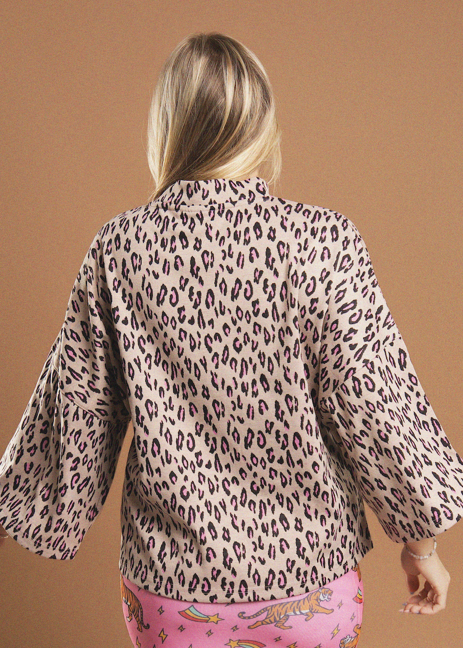 A woman shown from behind wearing a beige long-sleeve blouse with a dark brown leopard print pattern, paired with pink patterned pants, against a solid tan background.