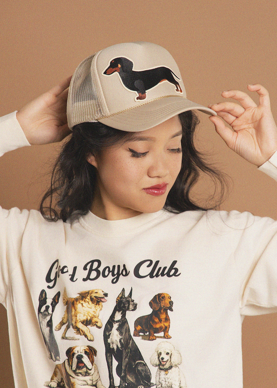 A woman wearing a beige trucker hat with a black dachshund graphic and a cream graphic sweatshirt that reads 'Good Boys Club' with dog illustrations, posed against a solid tan background.