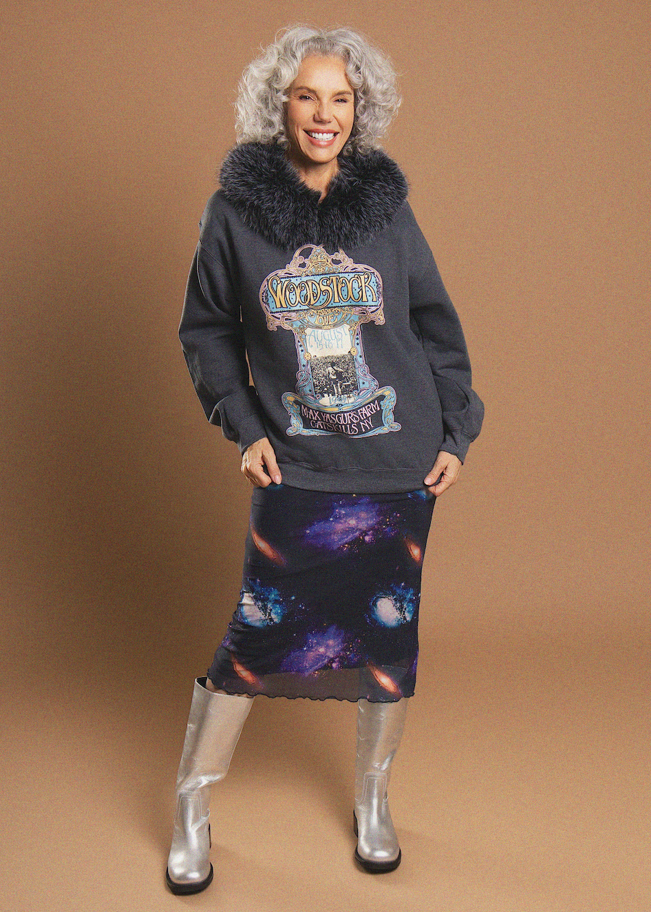 A woman modeling a dark charcoal gray sweatshirt with a black faux-fur collar and a large 'Wizards of the Coast' graphic on the front, paired with a dark galaxy-print skirt and silver knee-high boots, against a solid tan studio background.