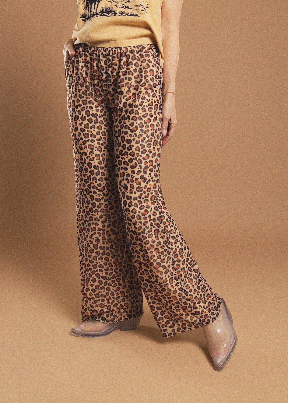 A model is shown from the waist down wearing leopard print wide-leg pants, a beige graphic t-shirt, and clear pointed-toe flat shoes against a solid tan studio background.
