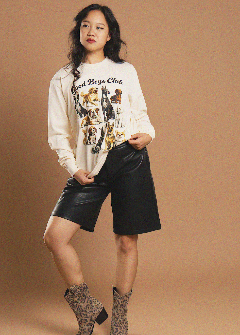A model posing in a cream-colored graphic sweatshirt featuring a dog print, paired with black faux-leather shorts and leopard-print ankle boots, set against a solid tan studio backdrop.
