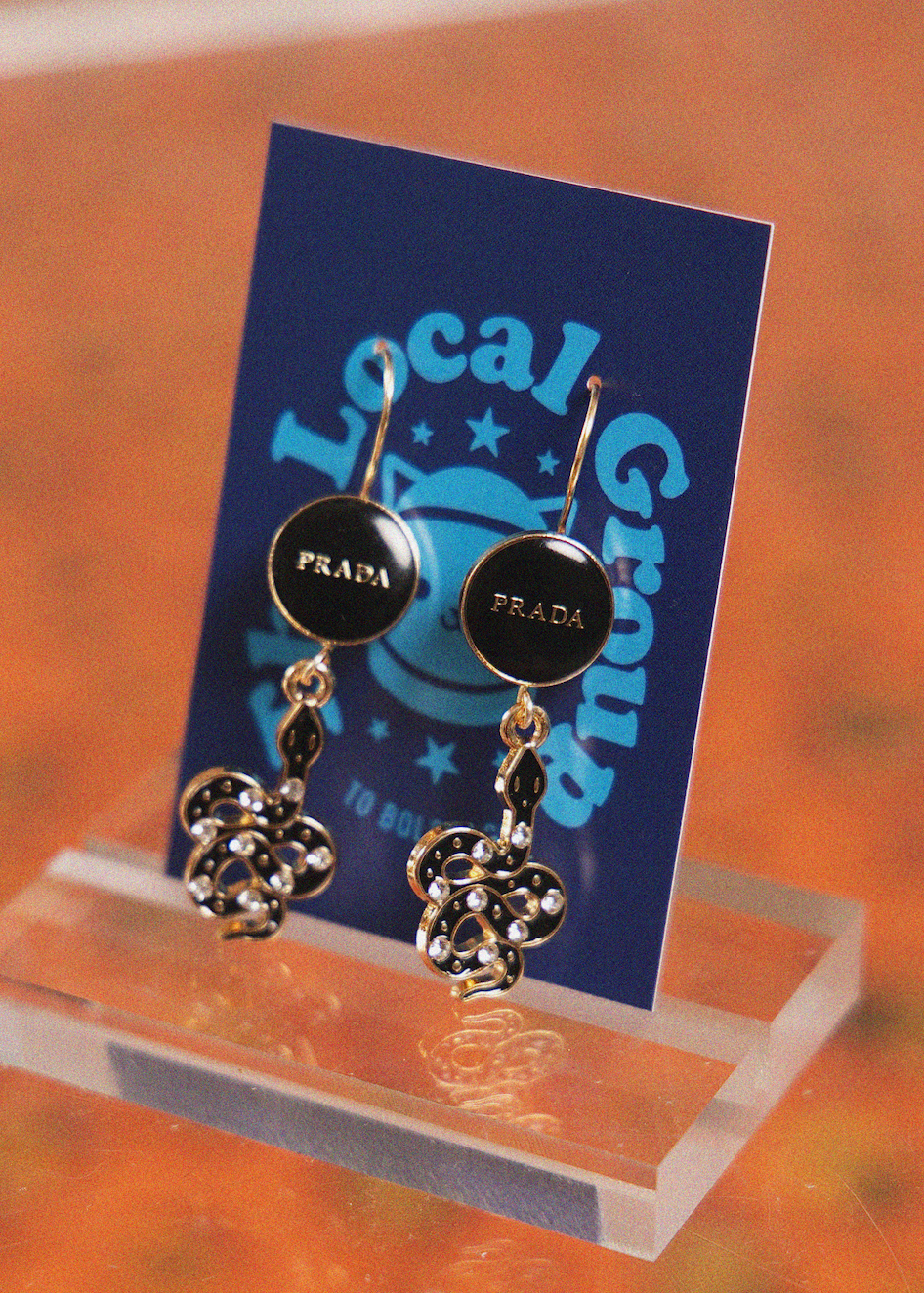 A pair of Prada drop earrings displayed on a blue Local Group retail card, resting on a clear acrylic stand against a warm, blurred orange-brown background.