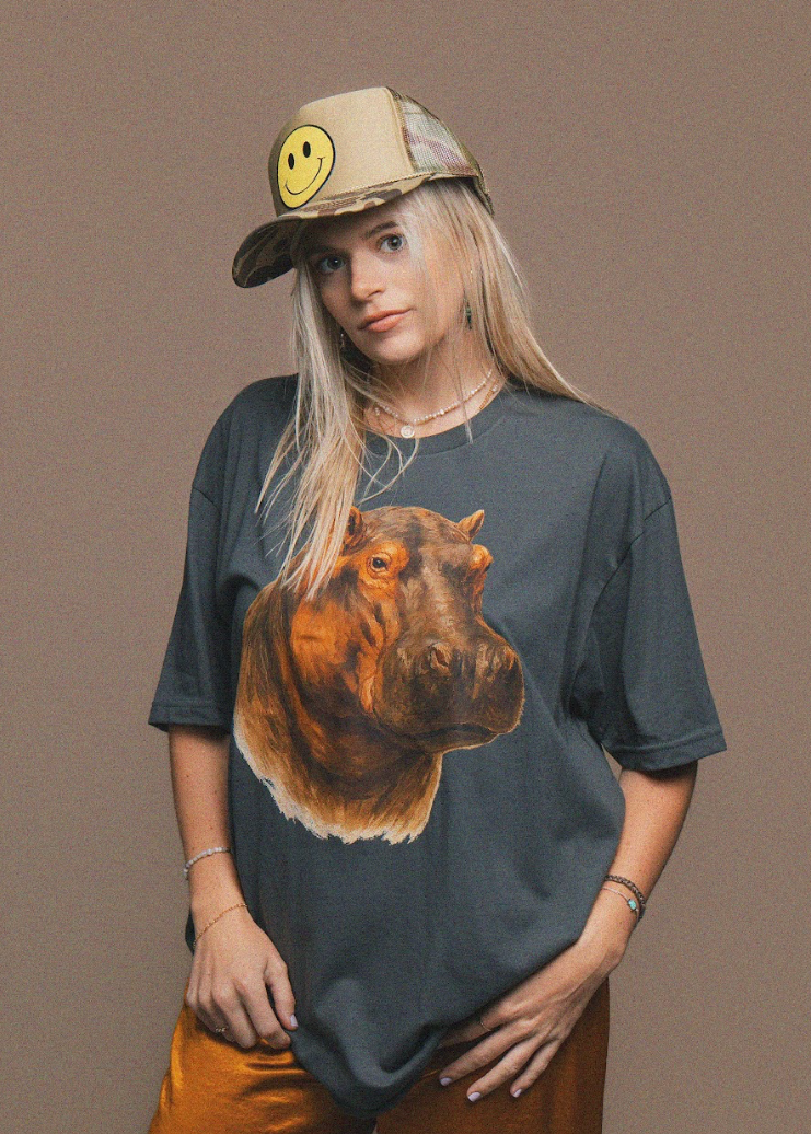Person wearing a t-shirt with a hippo design and a cap with a smiley face on a brown background