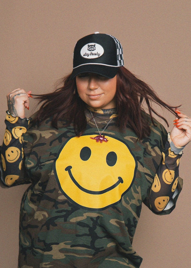 Person wearing a camouflage sweatshirt with a yellow smiley face and a black cap with white text.