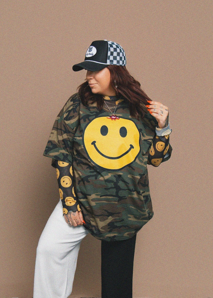 Person wearing a camouflage t-shirt with smiley face design and a checkered cap on a brown background